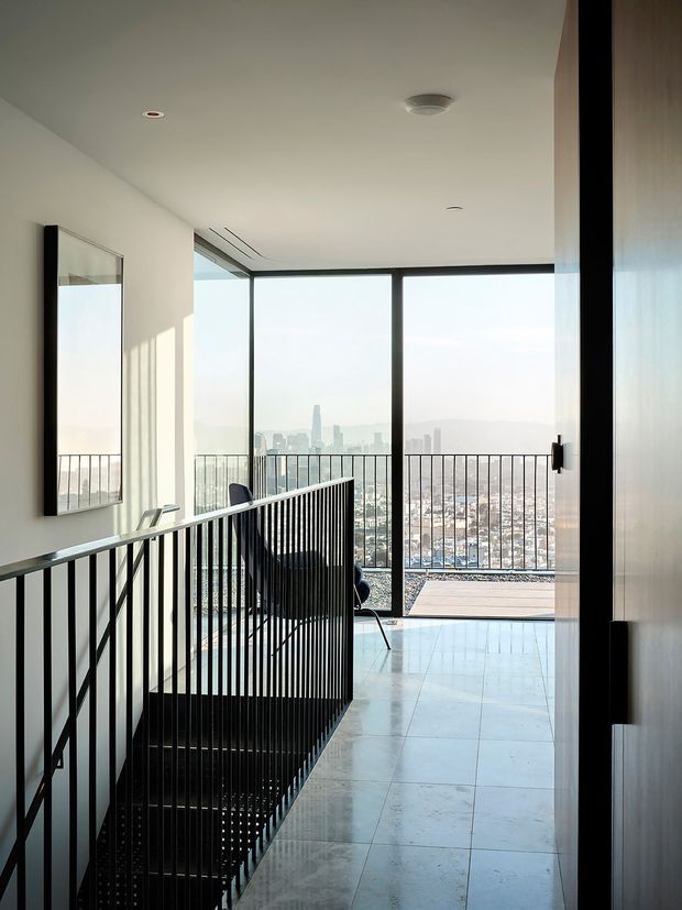 A bright, minimalist upper-level landing or hallway with a black vertical metal stair balustrade, pale large-format tiled flooring, white walls and ceiling, and expansive floor-to-ceiling glazing. The glazing opens onto a balcony with a matching black metal railing and sweeping urban skyline views. A tall framed mirror and discreet recessed lighting complete the refined contemporary interior.