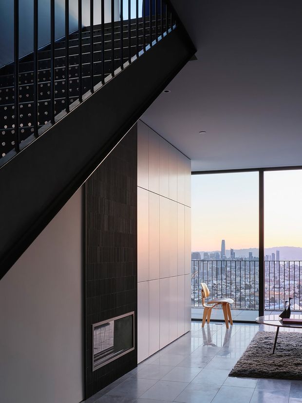 A dark, refined contemporary interior viewed beneath a dramatic black staircase with open metal balustrading. The space features a tall black tiled fireplace wall, pale full-height built-in wall panels, polished light flooring, and expansive floor-to-ceiling glazing opening to a balcony with a city skyline view. A small dining or reading setting and part of a living area are visible, creating a high-contrast, urban apartment atmosphere.