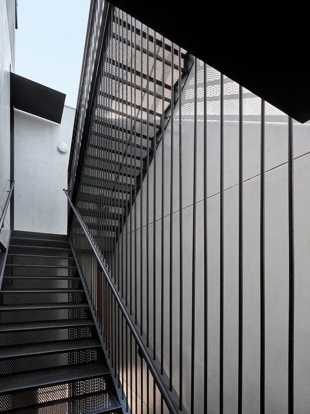 A narrow contemporary stairwell or circulation space with two flights of open-tread perforated metal stairs, dark metal handrails and balustrades, and a tall vertical metal screen. White rendered walls and overhead glazing or gridded screening bring in daylight, creating a stark industrial architectural composition.