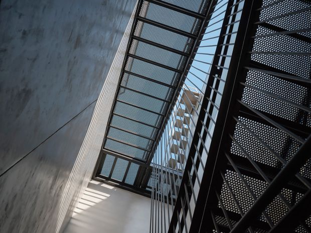 Upward-looking architectural view within a narrow, industrial-style atrium or stairwell. A black perforated metal stair or walkway structure rises along the right side, while a mesh-covered overhead bridge or skylight spans the space. Raw grey concrete walls, strong linear shadows, and glimpses of blue sky create a stark, dramatic composition.