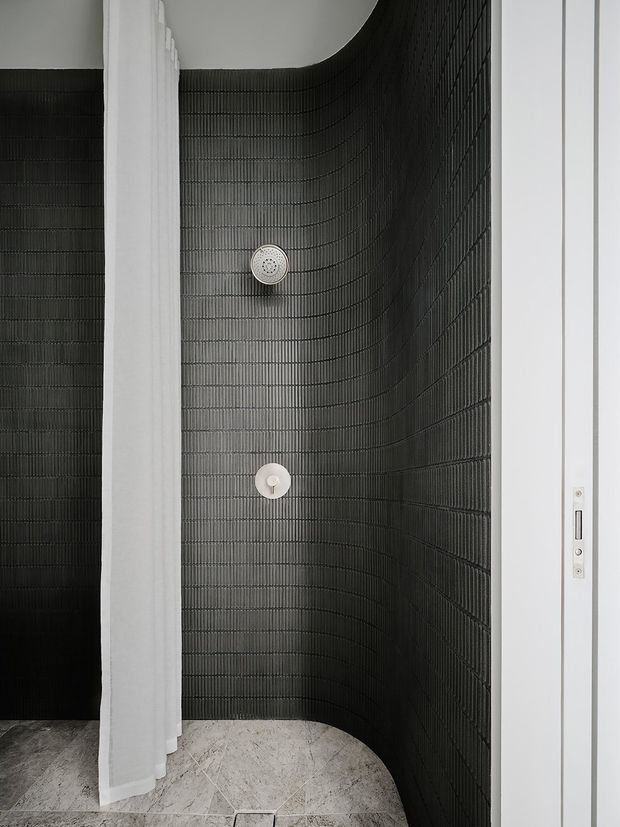 A minimalist walk-in shower enclosure with a curved wall fully clad in dark narrow-format ribbed tiles. A round overhead shower and matching control are set into the tiled wall, framed by crisp white walls and a white shower curtain. The floor is finished in pale stone-look tiles, creating a stark monochrome contrast. A slim white-framed door is visible at the right edge.