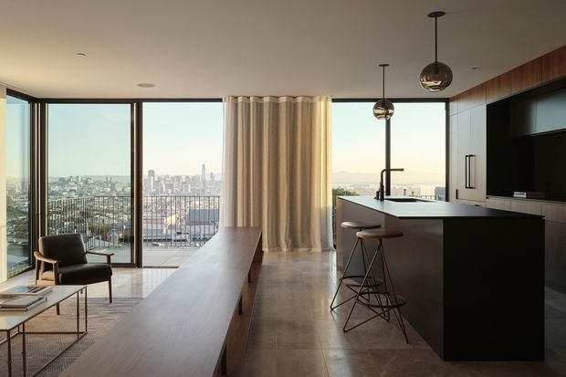 A contemporary open-plan kitchen and living area in a high-rise apartment, with floor-to-ceiling glazing and balcony views over a dense city skyline. The kitchen features dark built-in cabinetry, a large island with integrated sink and seating, warm timber wall cabinetry, polished stone-like surfaces, and suspended spherical pendant lights. A long timber console or dining bench extends through the living zone, which includes a lounge chair and coffee table. Neutral curtains and large-format flooring create a refined, warm modern interior.