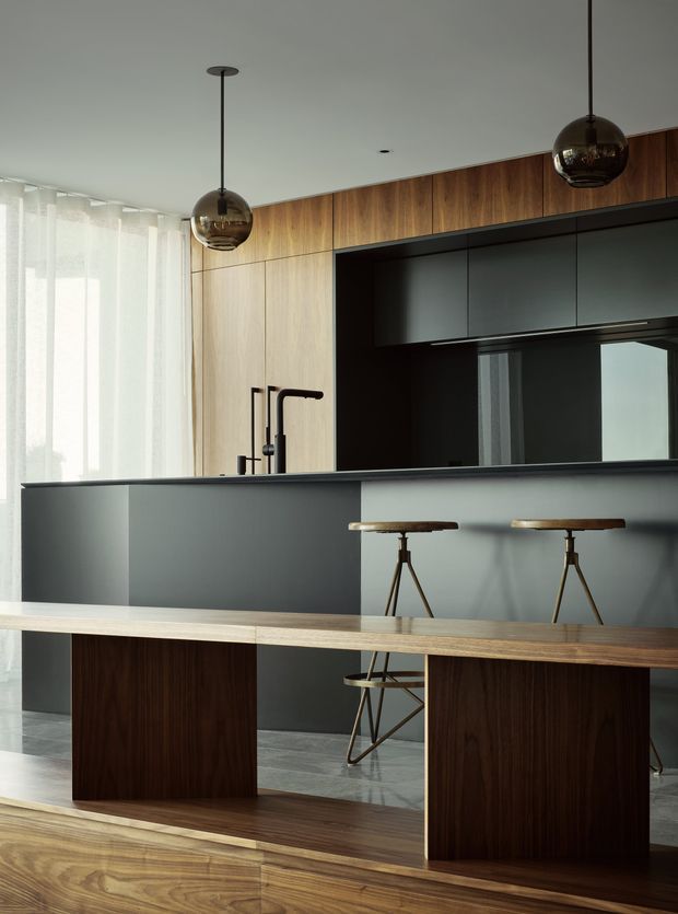 A refined modern kitchen with a dark charcoal island and integrated cabinetry, warm wood wall cupboards and timber dining or breakfast counter, and black pendant globe lights. A large window with sheer curtains provides soft daylight, while stools and a dark reflective splashback add depth to the compact open-plan space.