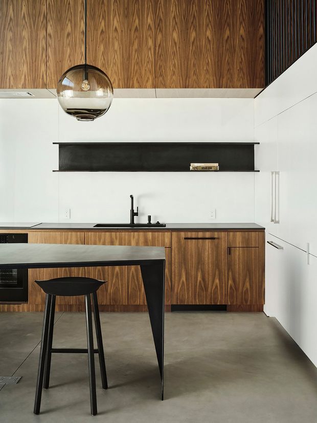 A refined modern kitchen featuring richly grained dark timber cabinetry and matching timber ceiling panels, white walls, a long black rangehood, black tapware, and a sculptural dark kitchen island with a black bar stool. The space has a polished yet restrained material palette, concrete flooring, integrated white cabinetry, and a smoked glass pendant light.
