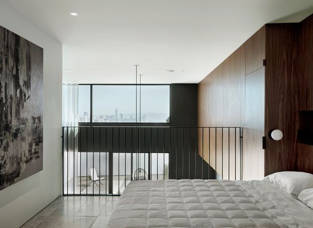 Contemporary bedroom overlooking a double-height void, with a low-profile bed in the foreground, full-height glazing, black metal balustrading, and a dramatic dark timber feature wall. A lower-level seating area is visible beyond the railing, while a large monochrome artwork adds contrast to the otherwise restrained interior.