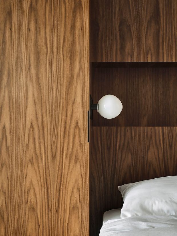Close-up interior view of a bedroom featuring a warm natural timber built-in wardrobe or door beside a dark timber wall and shelving unit. A small spherical wall light is mounted within the timber joinery, and part of a neatly made bed is visible at the lower right. The composition highlights the contrasting light and dark wood grain, concealed joinery, and minimal contemporary detailing.