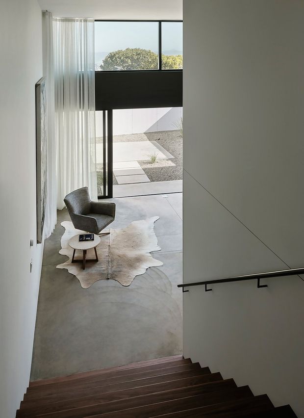 View down a minimalist interior stairway into a double-height living area. Dark timber stairs with a simple black handrail lead to a polished concrete floor furnished with a sculptural grey armchair, small round timber coffee table, and pale hide rug. Full-height glazing and sheer curtains open toward a geometric gravel courtyard, while white walls and dark window framing create a clean, high-contrast composition.