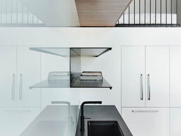 A minimalist contemporary kitchen viewed close-up, featuring extensive white flat-panel cabinetry with long vertical metal handles, a dark stone island benchtop with an undermount black sink and matte black gooseneck tap, and a floating open shelf displaying books. A pale timber ceiling panel and black-framed glazed balustrade are visible above, suggesting a mezzanine or stair opening.
