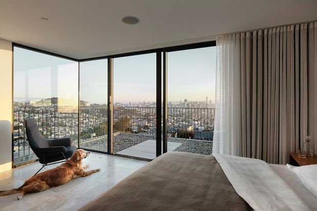 A contemporary bedroom with floor-to-ceiling corner glazing opening onto a balcony or terrace, overlooking a dense urban skyline. The room contains a bed with neutral bedding, a dark lounge chair, a small side table, and a dog resting on the polished pale floor. Full-height curtains frame the glazing, while black window frames and a simple recessed ceiling light create a clean modern interior.