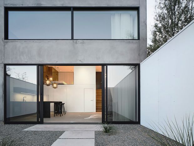 Exterior view of a minimalist two-storey concrete home with large black-framed glazed openings. The ground floor opens onto a gravel courtyard via expansive sliding glass doors, revealing a bright kitchen or living area with a compact island, pendant lighting, timber stair detailing, and a clean white interior. Concrete paving slabs lead through the gravel forecourt, with a tall white boundary wall and restrained planting.