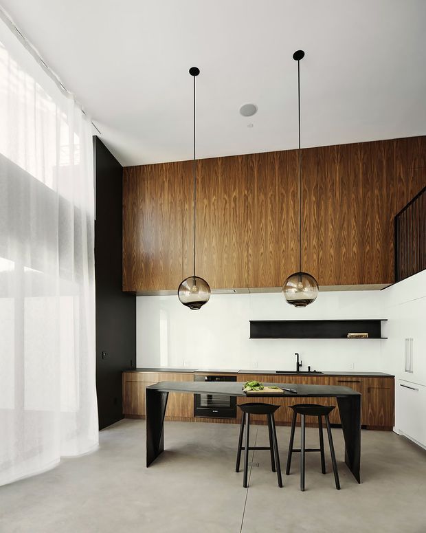 A contemporary kitchen with a dramatic full-height walnut-toned timber feature wall and matching lower cabinetry. A dark stone island with black geometric supports is paired with two slim black stools, while two globe pendant lights hang above. White walls and ceiling, black accents, polished concrete flooring, sheer floor-to-ceiling curtains, and a partial stair railing complete the bright, refined open-plan space.