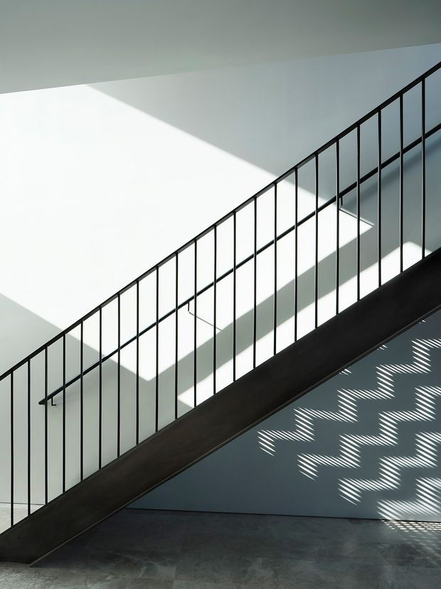 Minimalist architectural interior focused on a straight staircase with a dark solid stringer and slender black metal balustrade. Strong sunlight creates geometric shadows across pale walls and a dark tiled or concrete floor, emphasizing the clean, graphic composition.