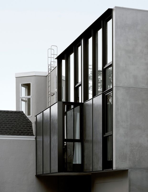 Photograph of a contemporary multi-storey building addition viewed from outside. The composition features a tall glazed facade with narrow vertical black window frames, dark metal cladding panels, and smooth light-grey rendered walls. A projecting upper-level volume forms a balcony or terrace edge, while an older rendered structure with a pitched dark shingle roof is visible alongside it. The design has a strong vertical rhythm and restrained monochrome palette.