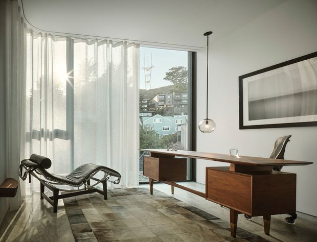 A refined home office or study with a mid-century-style wooden desk positioned beside full-height glazing and sheer curtains. The room includes a sculptural black leather chaise lounge, a framed monochrome artwork, a glass globe pendant light, and a patterned area rug. A residential neighbourhood is visible through the window.