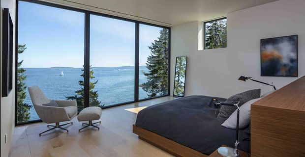 A contemporary bedroom with floor-to-ceiling corner glazing overlooking the ocean. The room features a low timber bed with dark charcoal bedding, a sculptural upholstered lounge chair and ottoman, a timber bedside or storage unit, minimalist lamps, pale timber flooring, white walls, and abstract wall art. A full-height mirror leans near the glazing, while coastal scenery and a sailboat are visible outside.