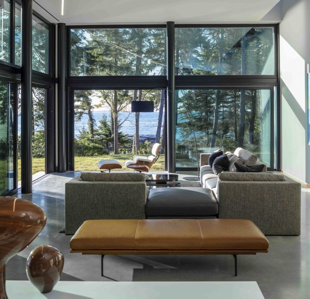 A sophisticated living room with expansive floor-to-ceiling glazing overlooking a wooded waterfront setting. The room features dark-framed windows, a polished concrete floor, low mid-century-inspired seating, a large brown leather bench, sculptural side tables, and a clean white ceiling. Strong natural light and the panoramic glazing create an open connection to the landscape.