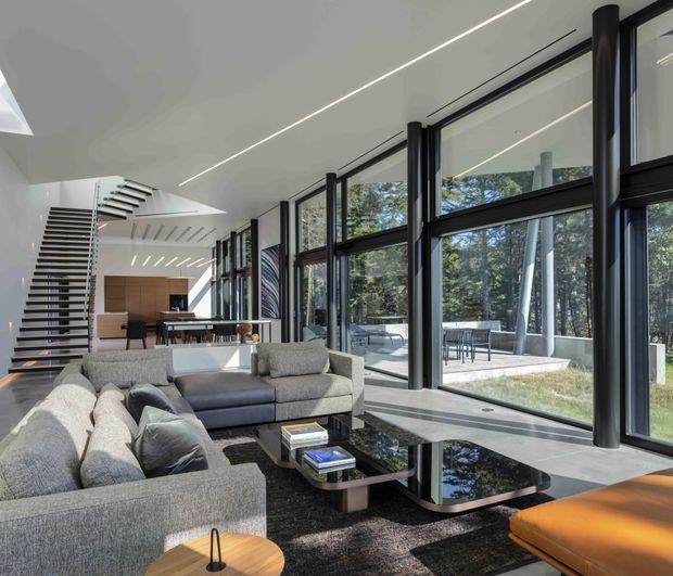 A spacious contemporary living room with large black-framed floor-to-ceiling glazing overlooking trees and an outdoor terrace. The room features low-profile grey sectional sofas, sculptural black glass coffee tables, a dark rug, a timber side table, and a dramatic open-riser staircase leading toward a dining and kitchen area. Clean linear ceiling lighting and exposed structural columns reinforce the modern architectural character.