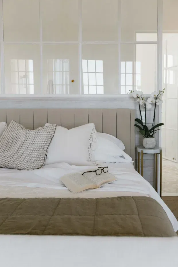 Bright, serene bedroom scene dominated by a neatly made bed with layered white bedding, a taupe quilted throw, textured neutral pillows, and a softly upholstered vertical-panel headboard. A small round side table holds a potted white orchid. The room features white-painted framed glass or mirrored partition panels and abundant reflected natural light, creating an airy, refined look.