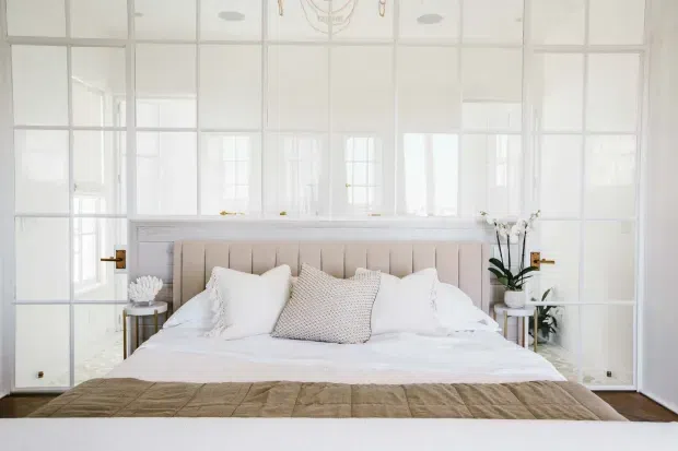 Bright, refined bedroom with a large upholstered bed centered against a pale paneled headboard. White-framed glazed partition walls and doors create an airy, light-filled backdrop, while layered white bedding, neutral textured cushions, a taupe quilt, slim bedside tables, brass-toned hardware, and potted orchids complete the serene interior.