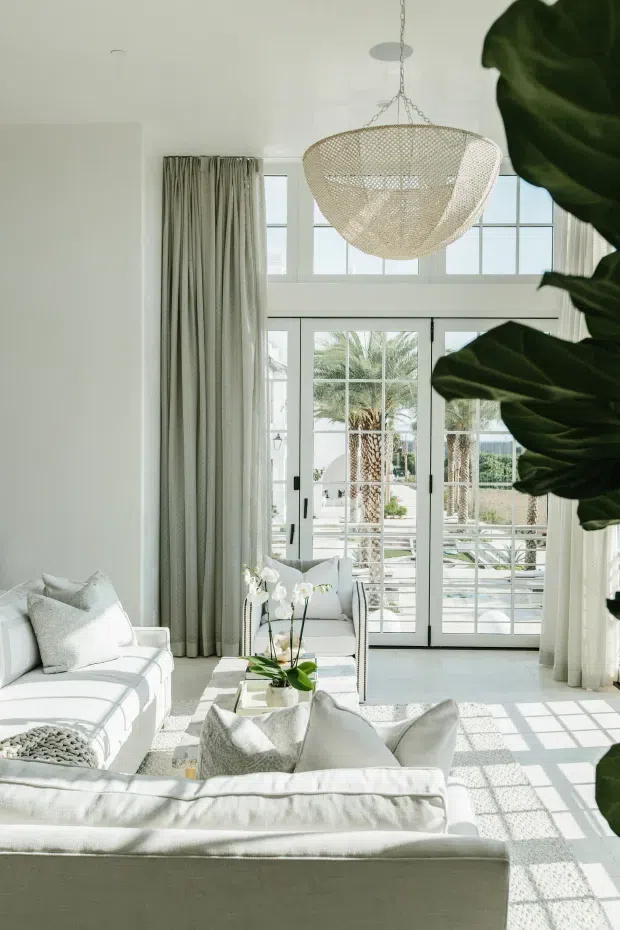 A bright, airy living room with tall white-framed French doors and clerestory windows opening to an outdoor terrace. The room features white upholstered sofas, pale neutral cushions, full-height light curtains, a glass coffee table with orchids, and a woven pendant light. Large leafy indoor plants frame the right side, while strong sunlight creates geometric shadows across the light floor.