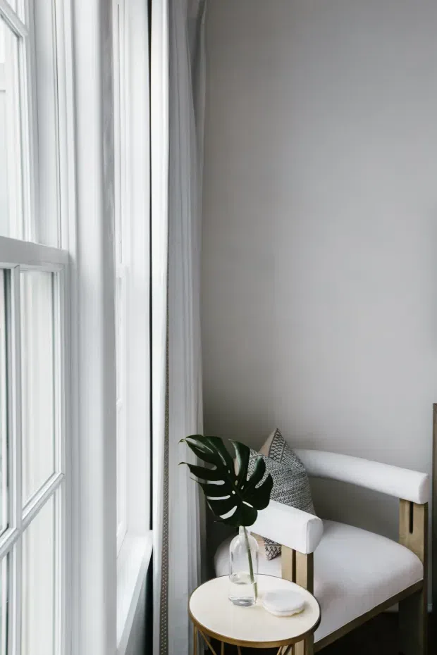 A bright, compact sitting nook beside tall white-framed windows. A white upholstered armchair with exposed light timber arms sits next to a small round light-coloured side table holding a glass vase with a large tropical leaf and a small dish. White curtains, pale walls, and soft neutral textiles create a clean, airy interior.