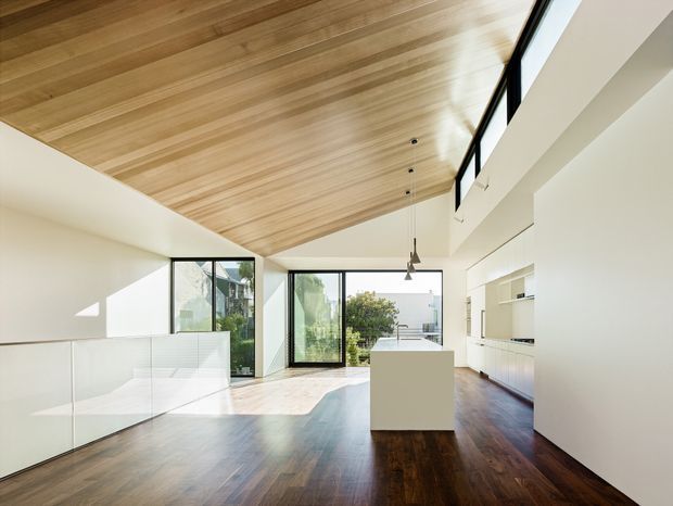 Bright, open-plan contemporary interior with a minimalist kitchen and living area. The space features a dramatic sloped timber-lined ceiling, dark timber flooring, white walls and cabinetry, a central white island, floor-to-ceiling black-framed glazing, and a raised clerestory window band. Natural light fills the room, with views toward greenery and neighbouring buildings.
