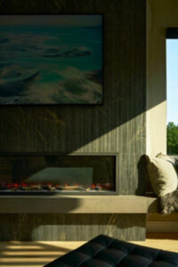 A moody living room detail featuring a dark vertically textured timber fireplace wall with a long linear firebox, a projecting stone or concrete hearth ledge, and a wall-mounted artwork above. Sunlight casts strong diagonal shadows across the feature wall and warm timber flooring. A dark tufted ottoman and part of a neutral upholstered chair are visible in the foreground and at right.