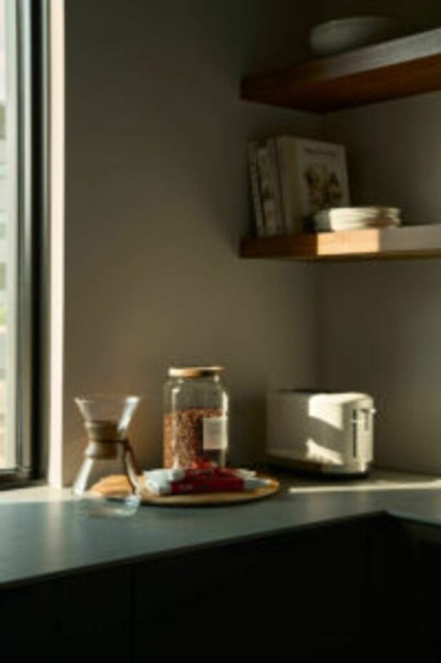A softly lit kitchen or pantry corner beside a window, featuring a dark countertop, dark lower cabinetry, warm wooden open shelves with books and dishes, and small countertop appliances and storage jars. Strong sunlight creates a calm, warm, intimate atmosphere.