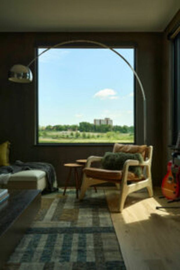 A warm, dark-toned bedroom or sitting area with a large square window overlooking open greenery and distant buildings. The room contains a low bed or daybed at left, a wooden-framed upholstered lounge chair, small side tables, a curved arc floor lamp, and a blue-grey patterned rug. Strong sunlight enters through the window, creating a relaxed mid-century-inspired atmosphere.