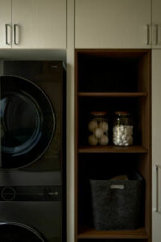Close-up view of a compact laundry area with stacked dark front-loading washer and dryer integrated into pale cabinetry. Beside the appliances is an open timber-toned shelving unit containing storage jars and a dark fabric basket. The cabinetry has simple vertical handles and a muted, functional built-in design.