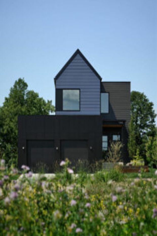 Exterior view of a contemporary dark-clad multi-level residence with a prominent front-facing gable, horizontal weatherboard cladding, large dark-framed windows, and an integrated two-car garage. The house is set behind landscaped planting under a clear sky.