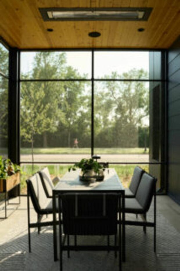 A compact dining area with a dark-framed floor-to-ceiling glazed wall overlooking trees and a lawn. A rectangular table with black framing and a pale top is surrounded by upholstered dining chairs. The room has a warm timber-lined ceiling with a recessed linear light and dark horizontal wall cladding, creating a simple indoor-outdoor connection.