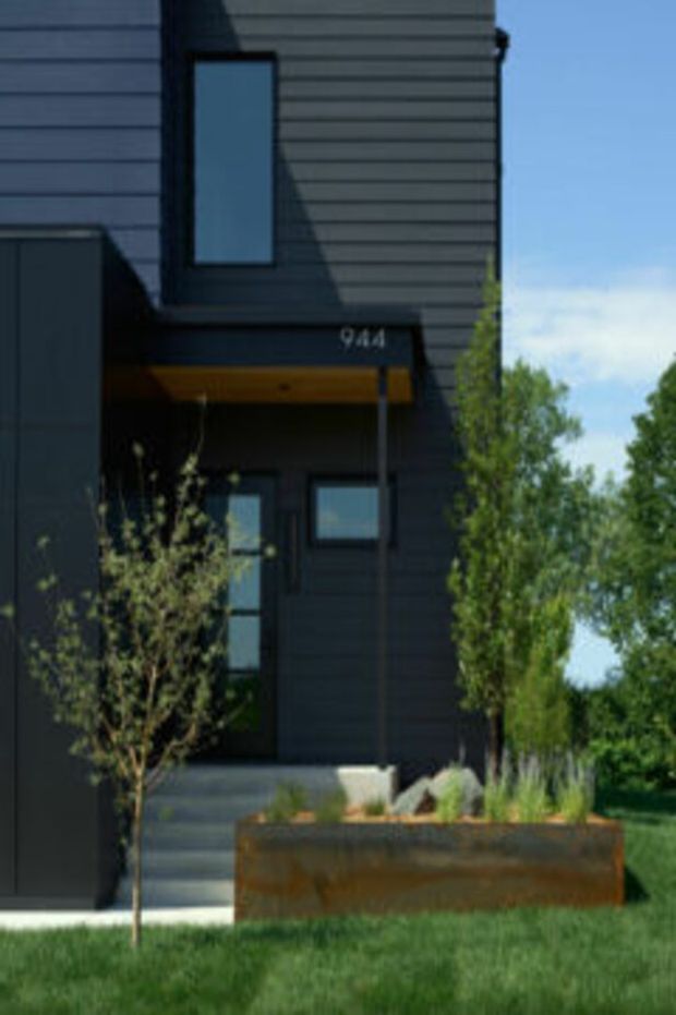 Exterior view of a narrow contemporary two-storey residence with dark horizontal cladding, black-framed windows, a recessed entry porch, concrete steps, and a rusted corten-steel-style raised planter in the foreground. The facade is set against a landscaped suburban garden with mature trees and lawn.