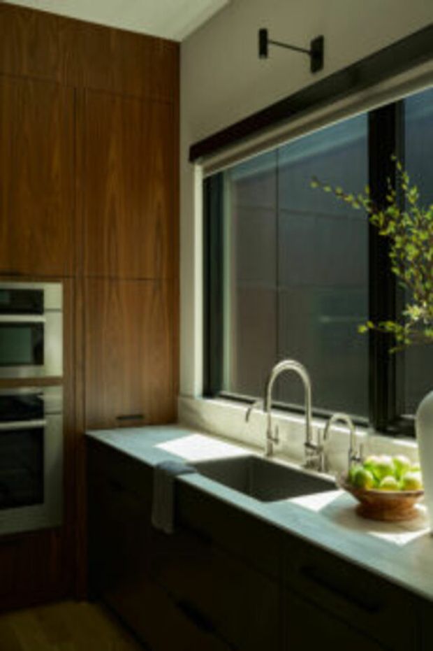 A modern kitchen focused on a long countertop beneath a large window. The space features dark stained timber cabinetry, integrated stainless-steel appliances, a pale stone benchtop, a deep undermount sink with a curved gooseneck tap, and dark window framing. Warm natural light enters through the glazing, creating a refined, contemporary atmosphere.