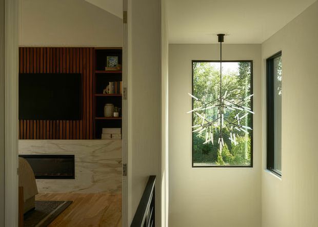 Interior view from a narrow circulation space toward a living room, with a dark-framed window overlooking greenery and a sculptural pendant light. The adjacent living area contains a large television set into a vertical timber-slat feature wall, open shelving, a pale stone fireplace surround, and timber flooring. The scheme is calm and refined with neutral walls, natural timber, stone, and black accents.