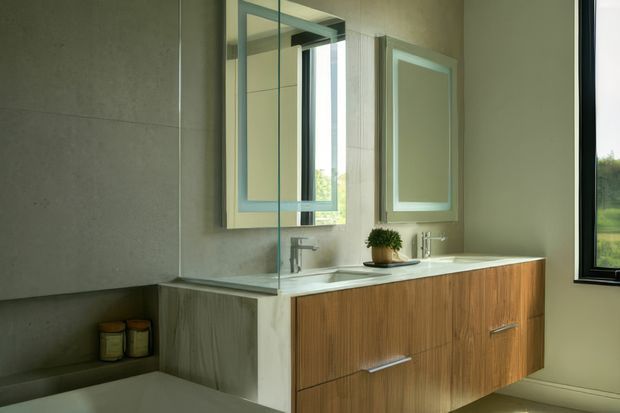 Contemporary bathroom vanity with a floating timber-look cabinet, pale stone countertop, twin wall-mounted mirrors, undermount basins and chrome tapware. Large-format grey wall tiles and a glass shower partition create a clean, refined interior, with natural light entering through the window.
