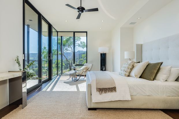 Bright, spacious contemporary bedroom with a large upholstered bed, neutral bedding, textured area rug, and minimal furnishings. Full-height black-framed glazed doors and windows open the room to a balcony or terrace with leafy landscape views. A sculptural ceiling fan, floor lamps, and a small desk or console complete the refined, airy interior.