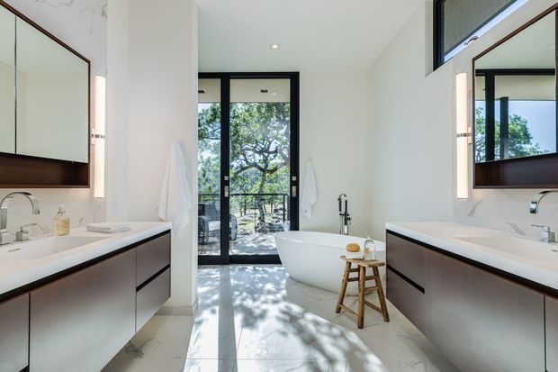 A bright, spacious contemporary bathroom with a freestanding white bathtub positioned between two large floating vanities. The room features white marble-look wall surfaces, pale stone or porcelain flooring, dark timber-toned vanity fronts, frameless mirrors with integrated vertical lighting, and black-framed glazed doors opening to leafy outdoor views. A separate shower area is visible through the glazing at the rear.