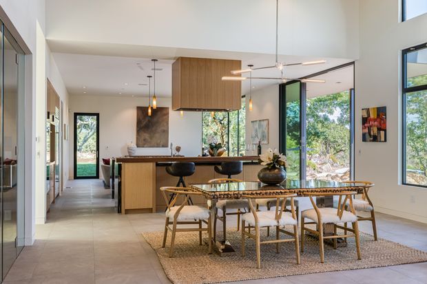 Open-plan contemporary kitchen and dining area with a high ceiling and large black-framed windows overlooking trees. The kitchen features a central island with warm timber cabinetry, a dark benchtop, pendant lighting, and bar stools. In the foreground, a light timber dining table with woven chairs sits on a textured neutral rug, while glazed doors and windows bring in abundant natural light.