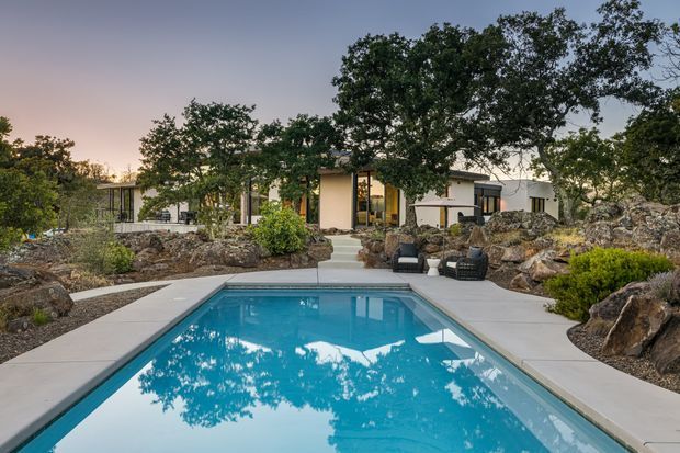 A modern single-storey residence viewed from the pool area at dusk. A rectangular turquoise swimming pool with pale concrete surrounds dominates the foreground, while a broad paved path leads toward the house. The home features low horizontal forms, extensive glazing, light rendered walls, dark-framed windows and doors, and a flat or very low-pitched roof. Natural rocky landscaping and mature trees surround the outdoor entertaining area.