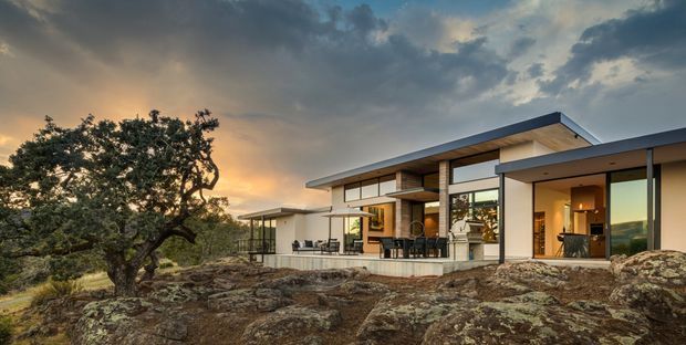 A low-slung contemporary single-storey house integrated into a rocky natural site. The exterior features clean horizontal roof planes, extensive floor-to-ceiling glazing, white rendered walls, dark metal roof and framing, and warm timber-lined soffits. Outdoor dining and lounge furniture sit on a terrace, while mature trees and exposed rock surround the home. The image is captured at sunset, creating a warm, dramatic atmosphere.