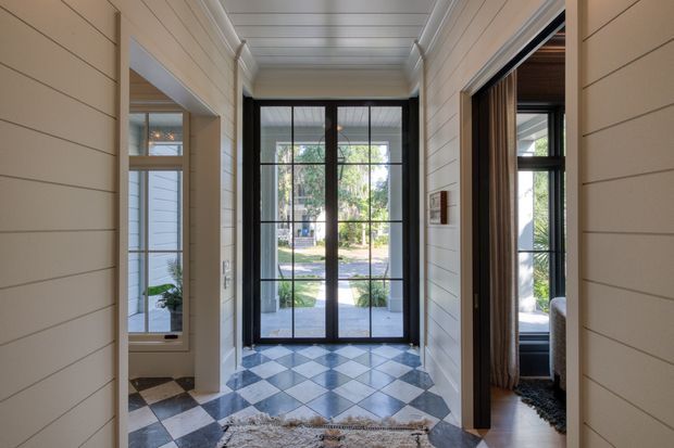 A bright, traditional-style residential entry hall with white horizontal wall panelling, a coffered or panelled ceiling, and a black-and-white checkerboard tile floor. A large black-framed glazed entry door opens toward a landscaped front yard, while open doorways reveal adjoining rooms on either side. The space is airy and symmetrical, with natural light, dark timber accents, and a small rug.
