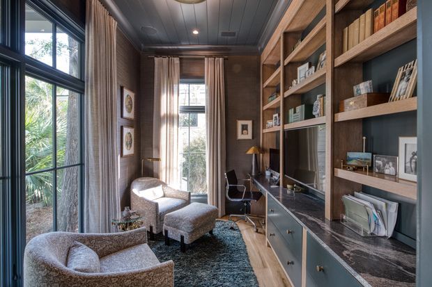 A refined, compact home office/library with floor-to-ceiling built-in timber shelving and cabinetry along one wall, incorporating a desk, drawers, open book storage, framed photographs, and a television or display screen. Two upholstered lounge chairs and an ottoman sit on a dark textured rug beside tall windows with full-height curtains. The room has dark timber wall and ceiling panelling, large black-framed glazing, warm layered lighting, and a sophisticated, intimate atmosphere.