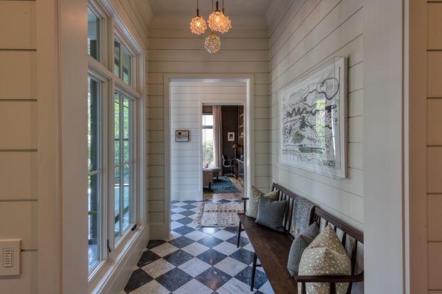 A narrow, light-filled traditional hallway or entry corridor with white horizontal wall panelling, tall multi-pane windows, and a striking black-and-white checkerboard tile floor. A dark wooden bench with patterned cushions lines the right wall beneath framed artwork, while decorative pendant lights hang overhead. An adjoining room is visible through the doorway at the end of the hall.