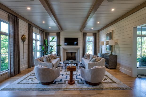 A bright, symmetrical living room with white horizontal wall panelling, exposed timber ceiling beams, tall divided-light windows, and a central fireplace with a mounted television above it. Two pairs of upholstered armchairs face one another around a glass coffee table, with a patterned area rug and a small central timber table. Dark curtains, indoor plants, and a textured console add warmth and contrast.