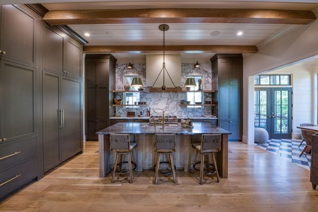 A spacious, traditionally styled kitchen centered on a large island with three stools. The room features dark grey built-in cabinetry, a marble-look backsplash, a substantial timber island, wide timber flooring, exposed timber ceiling beams, and a central pendant light. Glazed doors open to a bright adjoining space with patterned flooring.