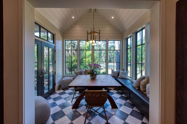 A bright enclosed sunroom or dining space with a vaulted, white-painted timber-lined ceiling, extensive black-framed glazing, and glazed double doors opening toward greenery. The room contains a long timber dining table, mixed dining chairs, and built-in bench seating with cushions. A striking checkerboard floor in alternating light and dark square tiles creates a graphic focal point, while a central pendant chandelier adds a refined traditional touch.