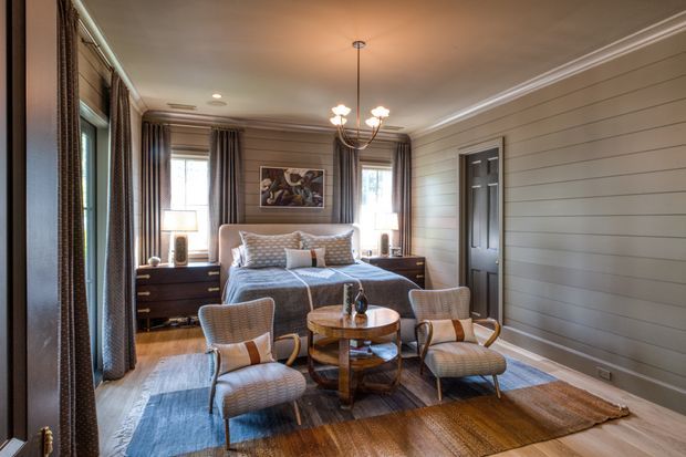 A warm, traditionally styled bedroom with a central bed, matching dark bedside cabinetry, upholstered and woven lounge chairs, a round timber coffee table, layered rugs, tall curtained windows, and horizontal wall panelling. Decorative crown moulding, a multi-arm pendant light, and a dark paneled door add refined architectural detail.