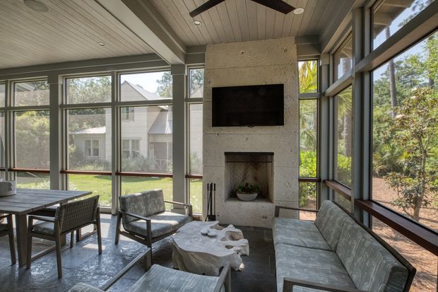 A bright, glazed living room or sunroom with floor-to-ceiling windows on multiple sides, a concrete fireplace wall with built-in television, a timber-lined ceiling, and a seating arrangement around a sculptural coffee table. The room opens visually onto a leafy garden and neighbouring residential setting.