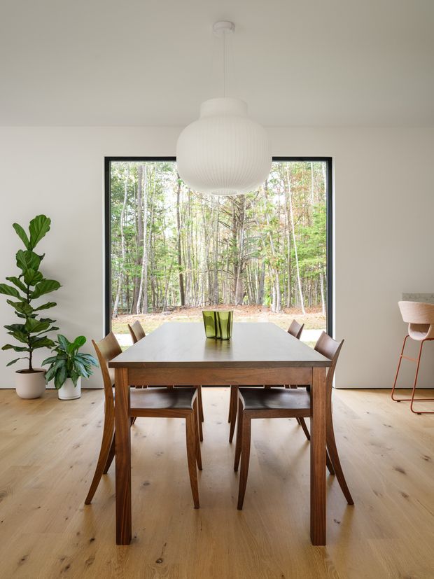 Bright, minimalist dining room with a large rectangular timber dining table and matching wooden chairs arranged in front of a full-width black-framed window overlooking woodland. Pale timber flooring and off-white walls create a warm, natural setting, complemented by indoor plants, a simple green glass centrepiece, and a large white sculptural pendant light. A bar stool is visible at the right edge.